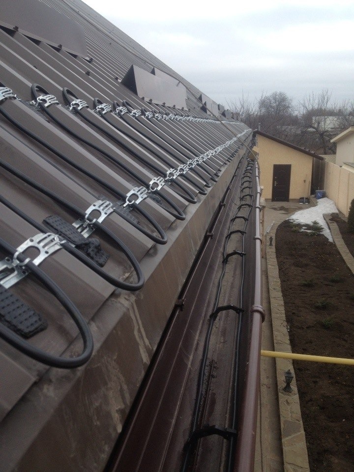 Brązowy dach z blachy trapezowej z zamontowanymi przewodami grzejnymi w rynnie i na połaci, zabezpieczonymi metalowymi uchwytami, widoczne zanieczyszczenia na blasze.