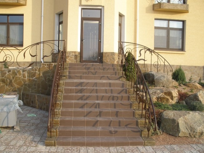 Zewnętrzne schody wejściowe wyłożone brązowymi płytkami, ozdobione kutymi, brązowymi balustradami z motywem roślinnym, prowadzące do drzwi wejściowych budynku z elewacją w kolorze ciepłego beżu.