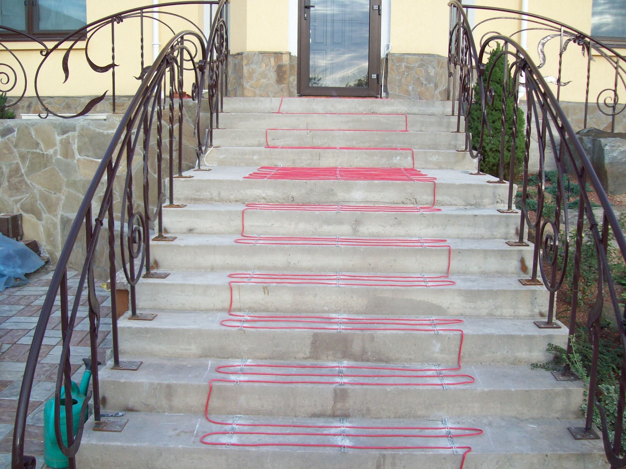 Betonowe schody wejściowe z zamontowanym systemem ogrzewania kablowego, czerwone przewody ułożone równolegle na stopniach, metalowa balustrada z ozdobnymi elementami.