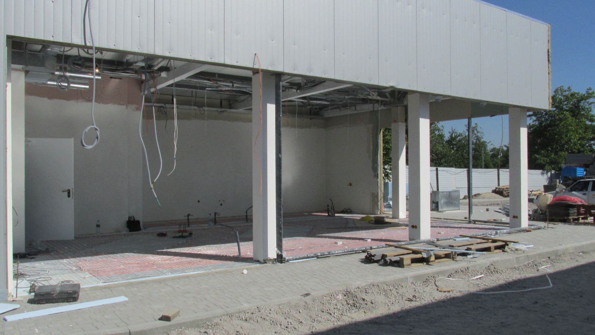 Montaż czerwonych kabli grzejnych na podłożu wewnątrz budynku stacji benzynowej w trakcie budowy, widoczne kable zwisające z sufitu, białe kolumny i ceglana kostka na zewnątrz.