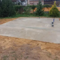 Świeżo wylany fundament betonowy z widocznymi pionowymi rurami instalacyjnymi, otoczony piaskiem i ziemią, w tle zieleń i częściowo widoczny budynek w trakcie budowy.