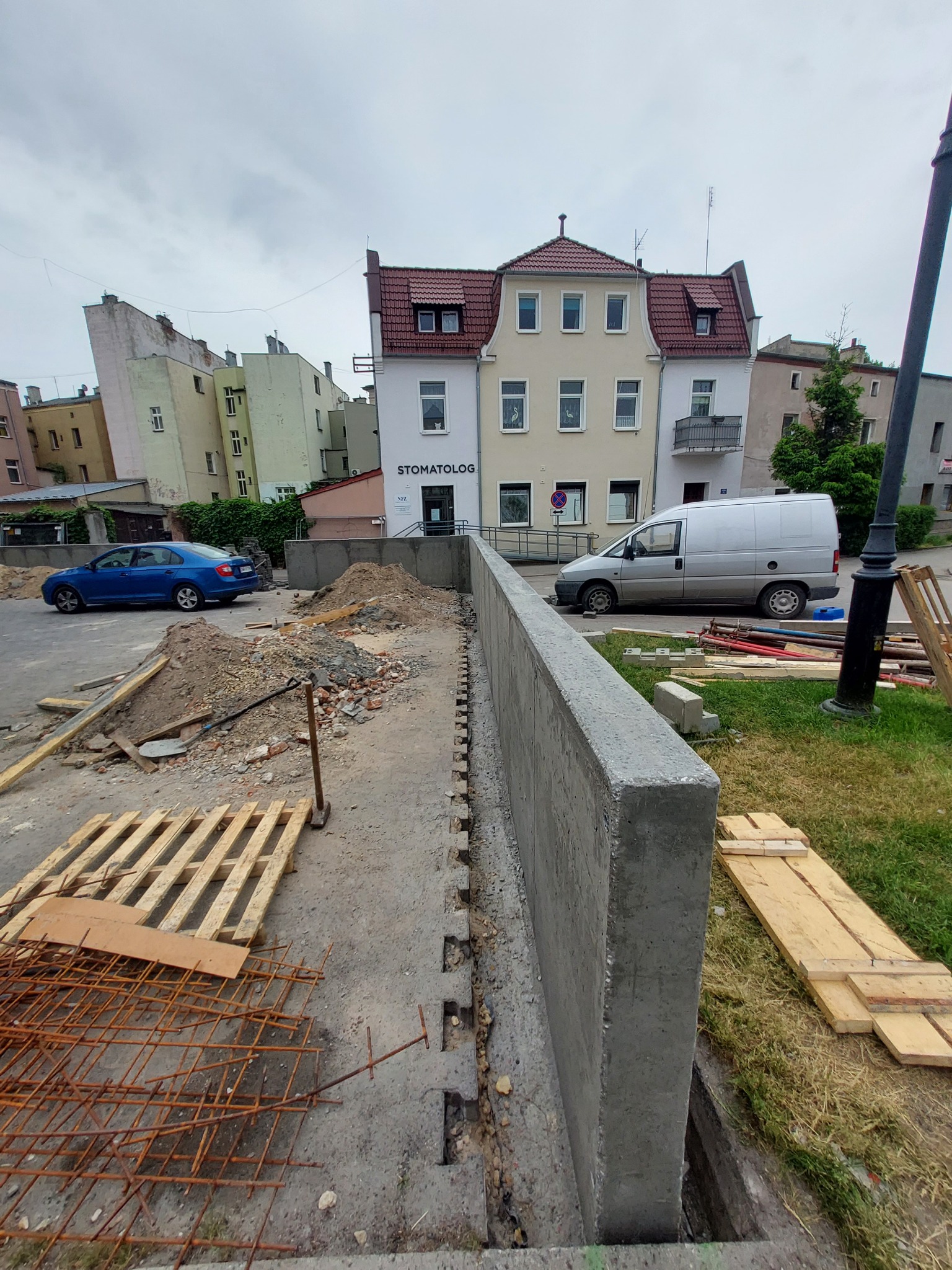 Wznoszenie betonowej ściany oporowej przy ulicy w zabudowie miejskiej, widoczne zbrojenie i deskowanie, w tle budynki mieszkalne i samochody.