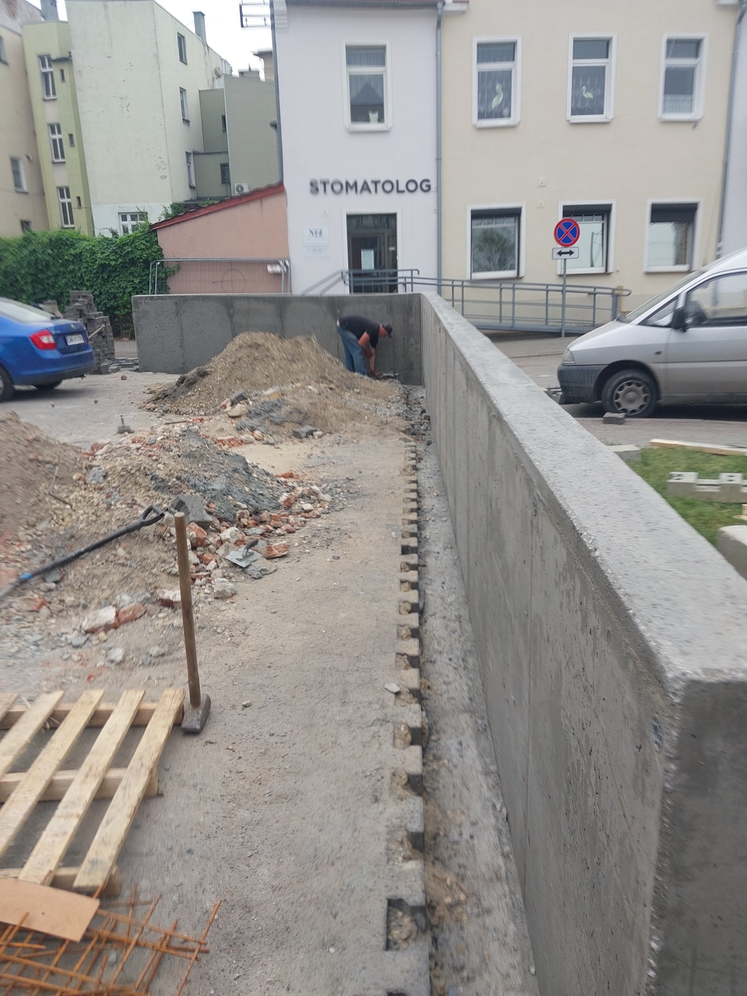 Betonowy murek oporowy w trakcie budowy, widoczne ślady po szalunku, obok stosy ziemi i gruzu, w tle budynek z napisem 'Stomatolog' i zaparkowane samochody.