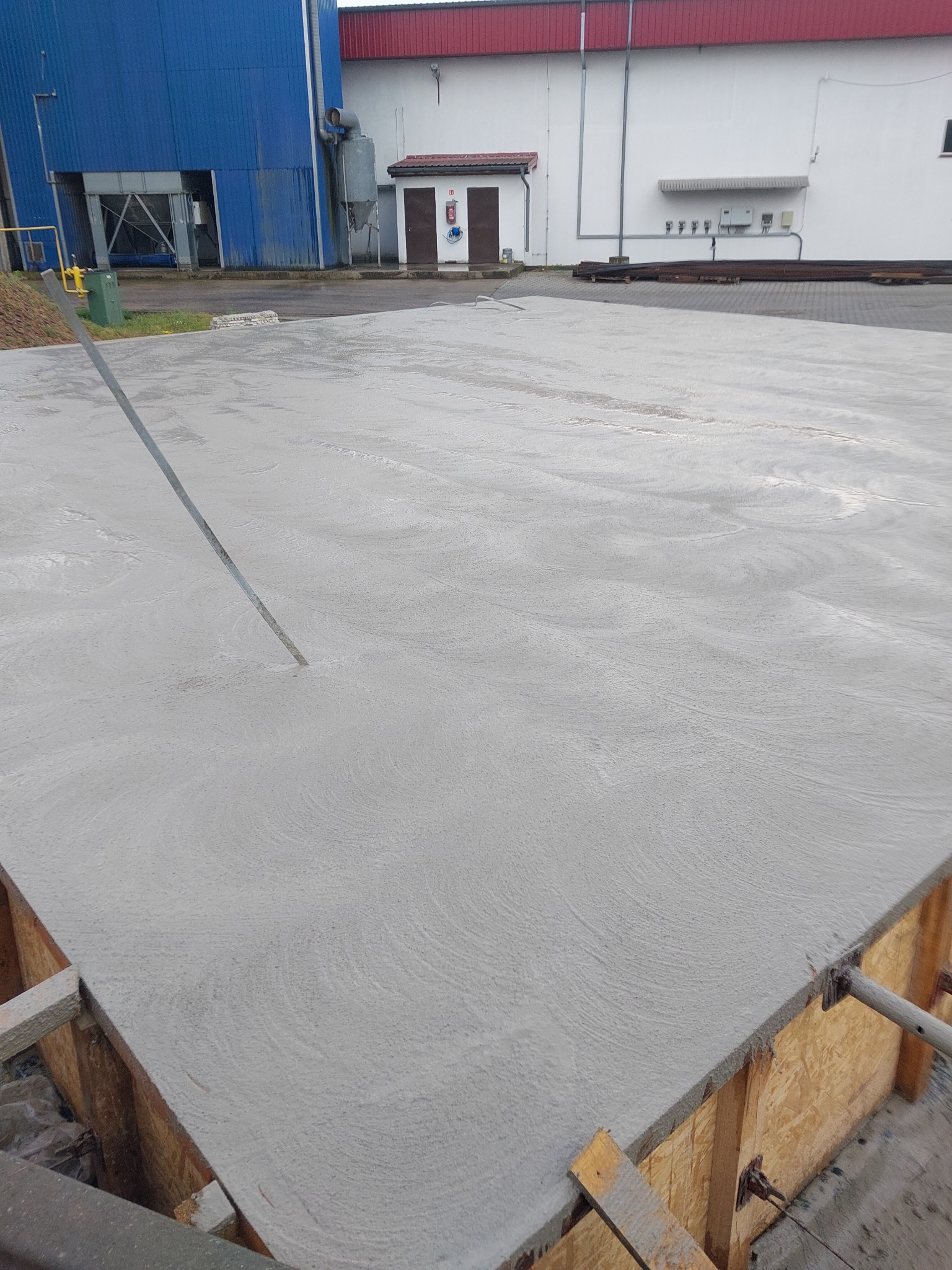 Świeżo wylana, gładka betonowa płyta w drewnianym szalunku, wyrównywana długą łatą w otoczeniu przemysłowym.