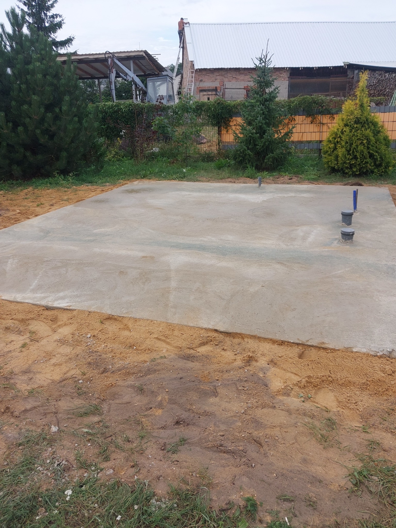 Świeżo wylany fundament betonowy z widocznymi pionowymi rurami instalacyjnymi, otoczony piaskiem i ziemią, w tle zieleń i częściowo widoczny budynek w trakcie budowy.