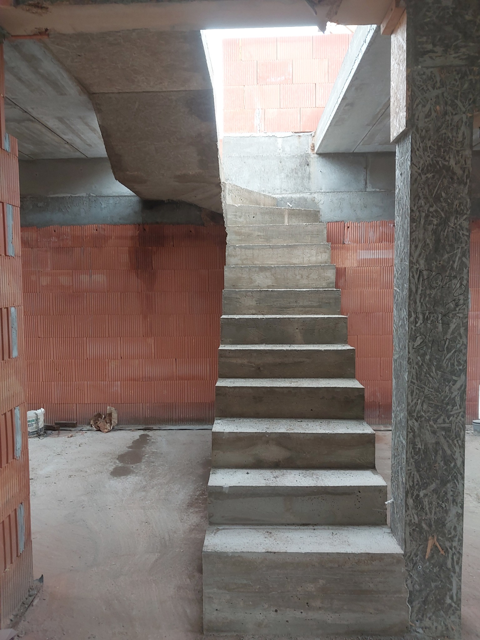 Surowe betonowe schody wewnętrzne w trakcie budowy, widoczne ślady po deskowaniu, murowane ściany z pustaków ceramicznych w tle, kolumna z płyty OSB po prawej stronie.
