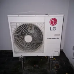 Zewnętrzna jednostka pompy ciepła LG Therma V na metalowych wspornikach, umieszczona przy szarej ścianie na ciemnej, mokrej kostce brukowej, widoczne opadłe liście.