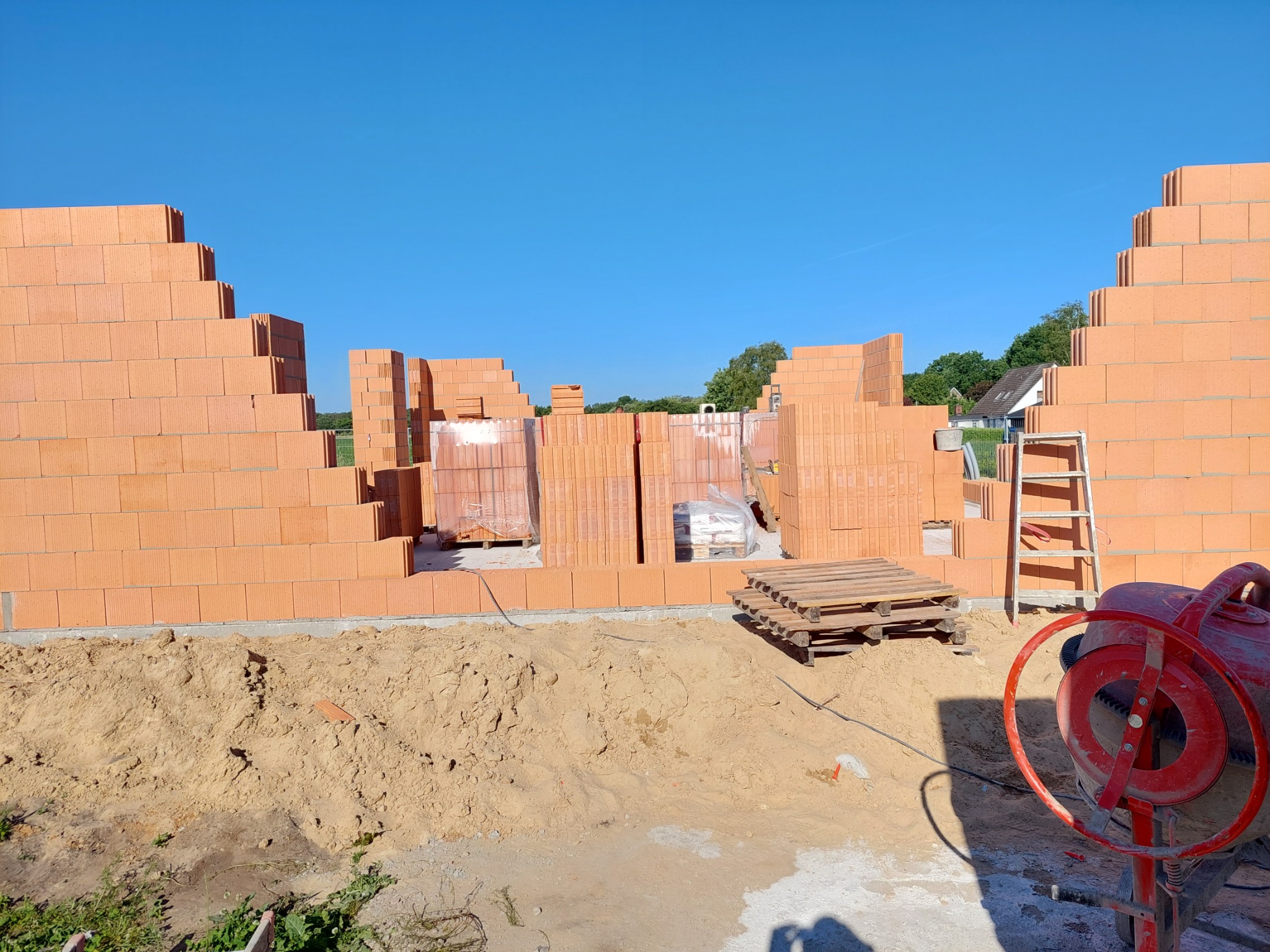 Wznoszone ściany budynku z bloczków ceramicznych, widoczna zaprawa, paleta z bloczkami, drabina i czerwona betoniarka na tle błękitnego nieba.