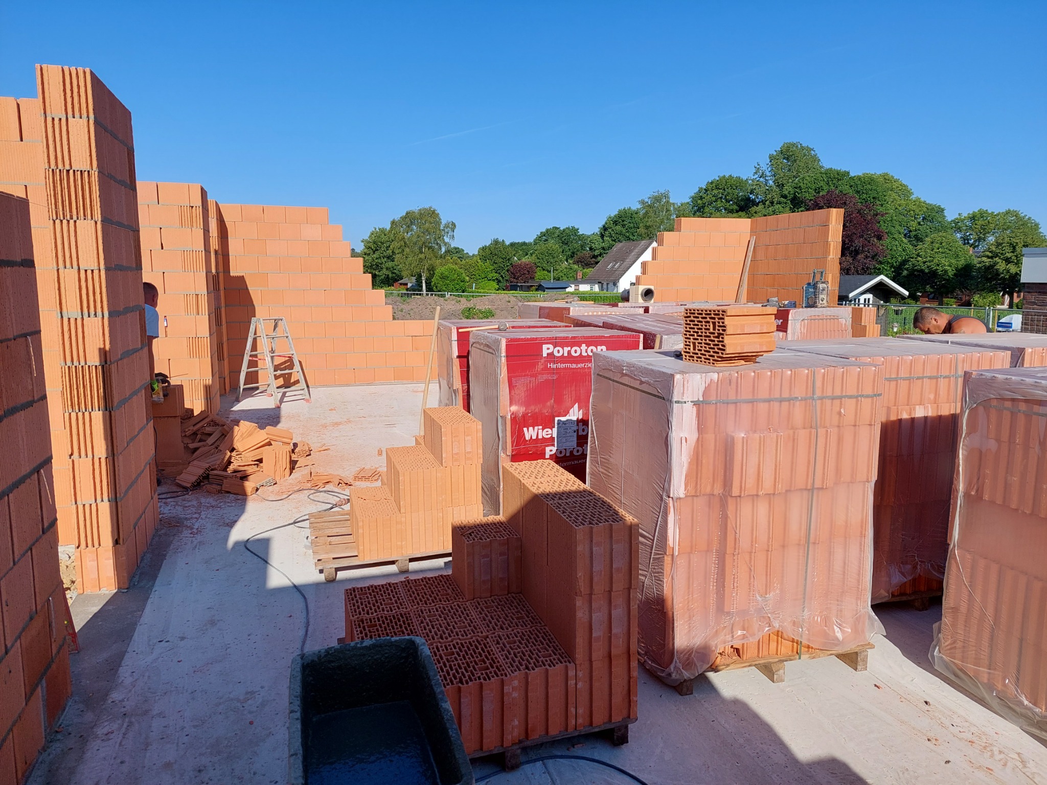 Wznoszenie ścian domu z czerwonych pustaków Poroton na zaprawie klejowej, widoczne palety z pustakami, drabina i pracownik w tle