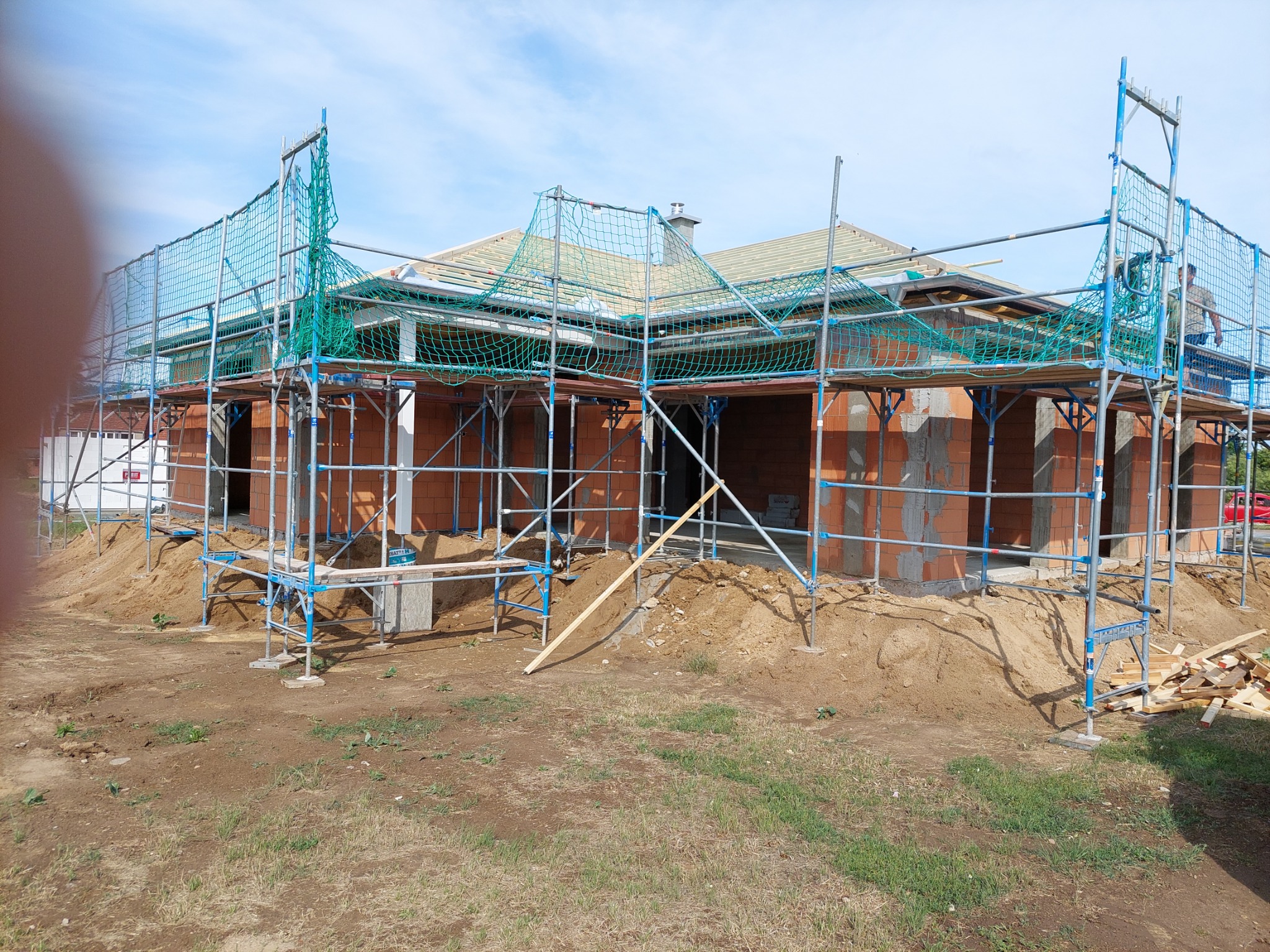Budowa domu jednorodzinnego w trakcie wznoszenia ścian z czerwonej cegły, z widocznym rusztowaniem zabezpieczonym siatką, drewnianą konstrukcją dachu i przygotowanym terenem.