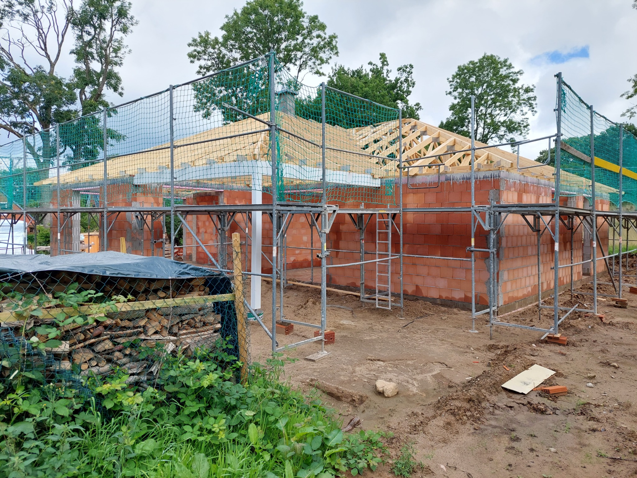 Wznoszenie ścian z czerwonej cegły na budowie domu, widoczne rusztowania, zabezpieczająca siatka i drewniana konstrukcja dachu w trakcie budowy.