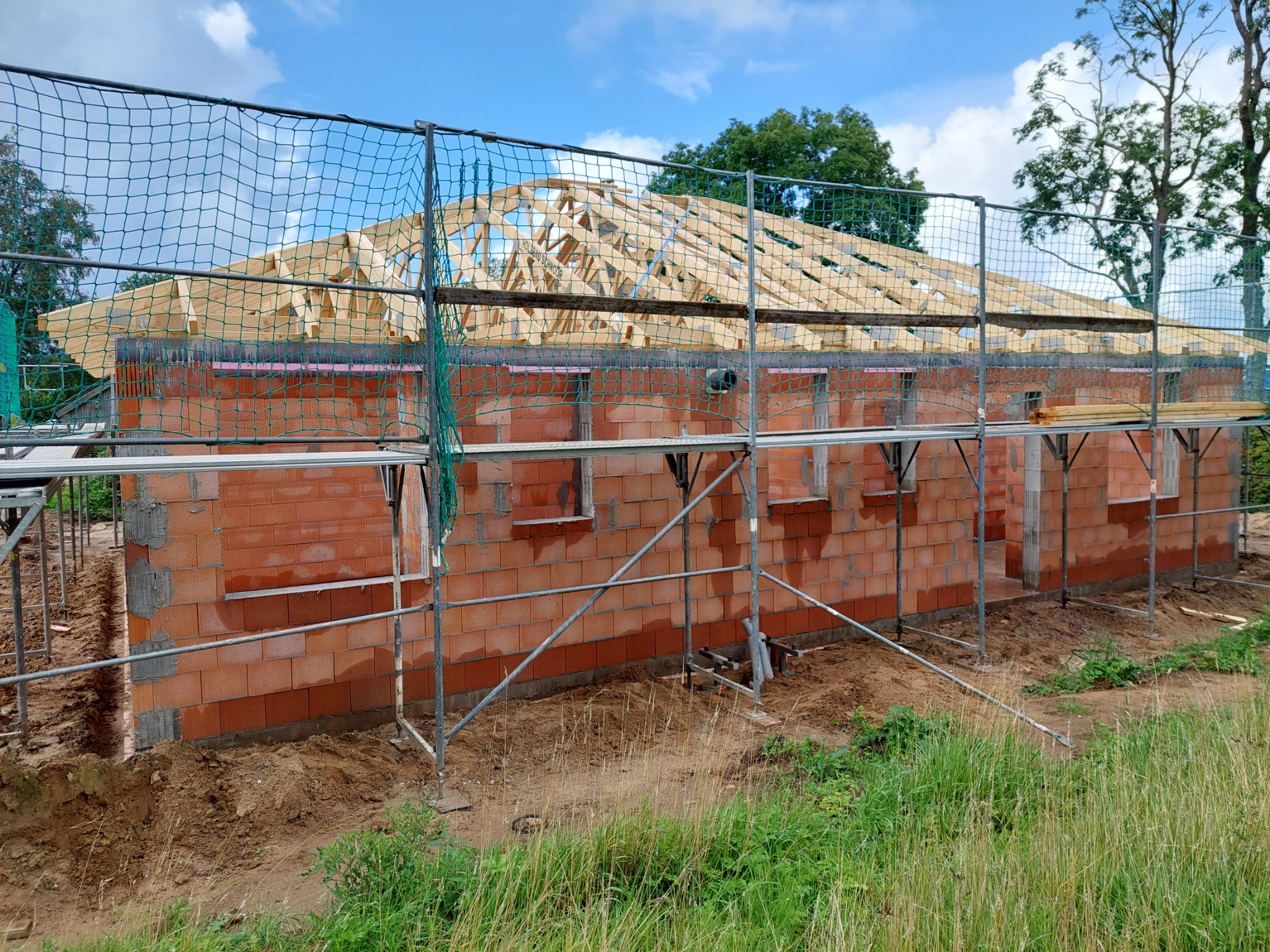 Wznoszona konstrukcja domu z czerwonej cegły, zabezpieczona siatką ochronną i rusztowaniem, z widoczną drewnianą konstrukcją dachu w trakcie budowy, na tle błękitnego nieba.