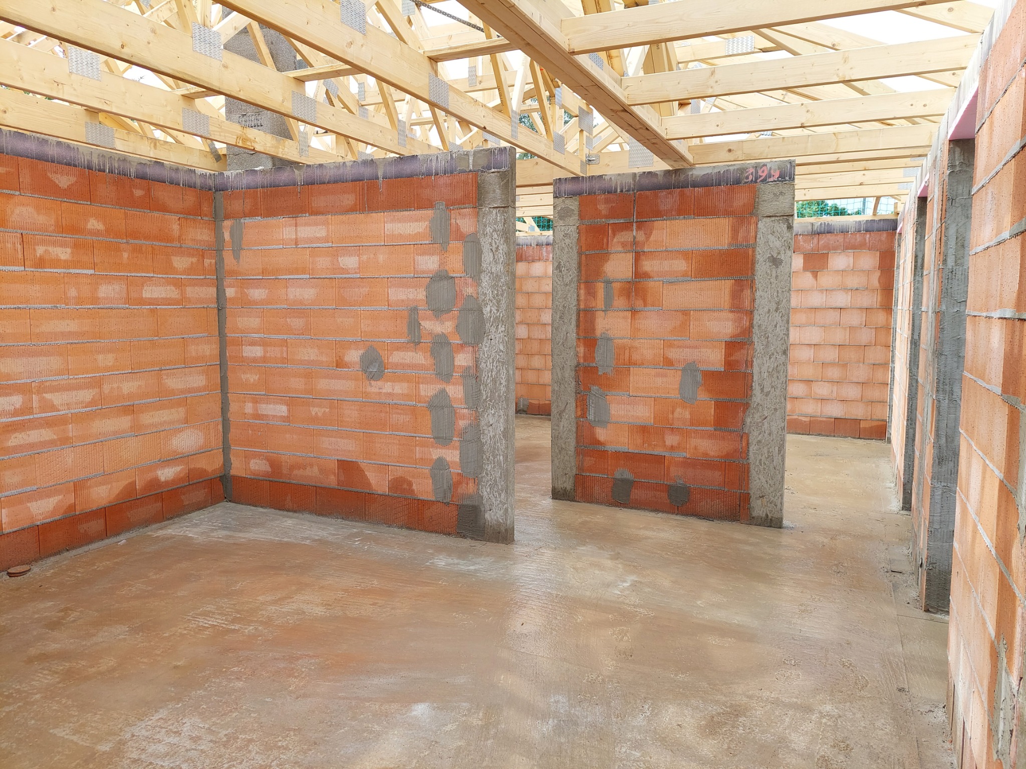 Wnętrze domu w trakcie budowy: murowane ściany z czerwonej cegły ceramicznej, betonowa wylewka podłogowa i drewniana konstrukcja dachu.
