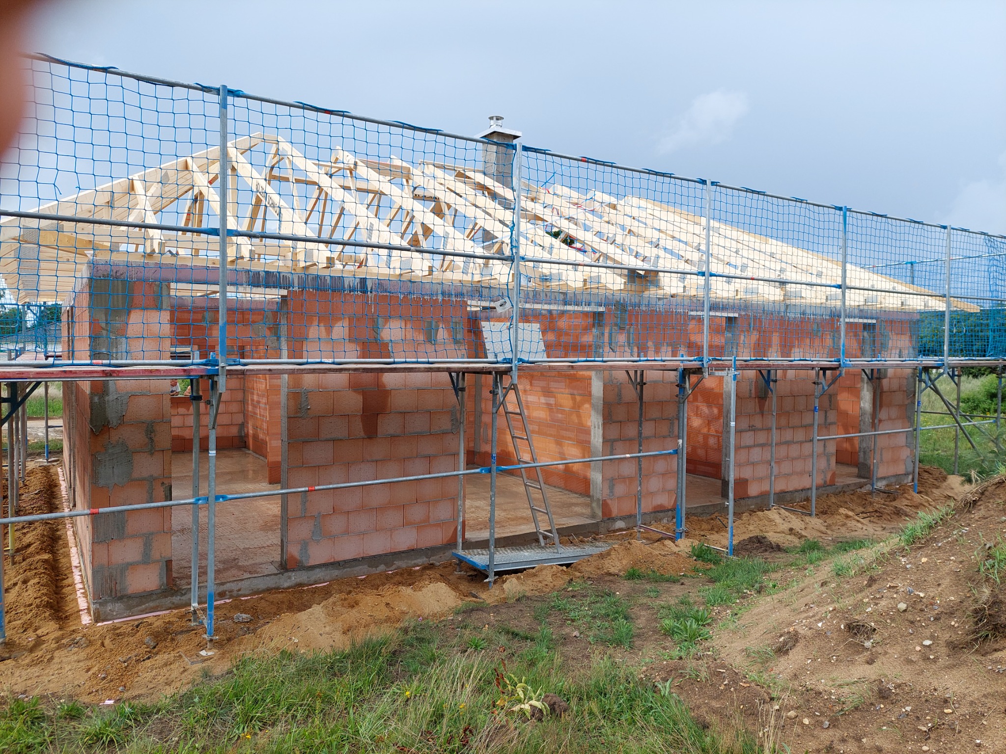 Surowy stan domu z czerwonej cegły z drewnianą konstrukcją dachu, zabezpieczony niebieską siatką rusztowaniową, widoczne rusztowania i drabina.