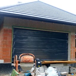 firmadavex.pl - Nowo budowany garaż z czarną segmentową bramą garażową, jeszcze w folii ochronnej, z pomarańczową betoniarką na pierwszym planie i stertą bloczków obok.