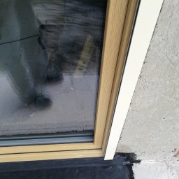 firmadavex.pl - Fragment okna PCV z drewnopodobną ramą i białą listwą wykończeniową, widoczne uszczelnienie czarną masą bitumiczną, refleksja postaci w szybie.