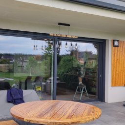 firmadavex.pl - Duże przesuwne okno tarasowe z czarnymi ramami, odbijające postać na drabinie, widok z zewnątrz, drewniany stół na pierwszym planie.