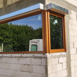 firmadavex.pl - Nowo zamontowane okno PCV z brązową ramą i metalową osłoną zewnętrzną na surowej ścianie budynku w trakcie budowy, z widocznym odbiciem otoczenia.