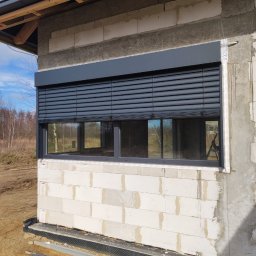 firmadavex.pl - Elewacja surowego budynku z zamontowanymi zewnętrznymi roletami w kolorze grafitowym, widok na okno z zamkniętymi lamelami i fragment krajobrazu w tle.