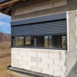 Elewacja surowego budynku z zamontowanymi zewnętrznymi roletami w kolorze grafitowym, widok na okno z zamkniętymi lamelami i fragment krajobrazu w tle.