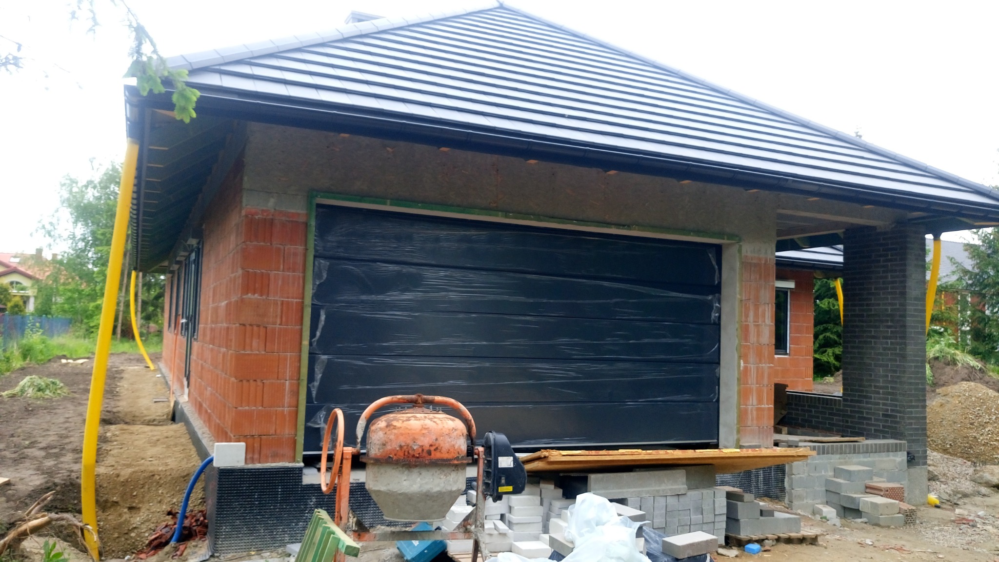 Nowo budowany garaż z czarną segmentową bramą garażową, jeszcze w folii ochronnej, z pomarańczową betoniarką na pierwszym planie i stertą bloczków obok.