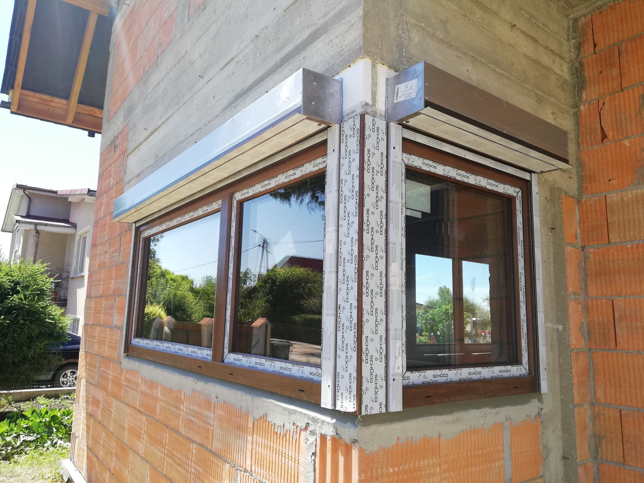 Surowy stan montażu okna PCV z roletą zewnętrzną w nowo budowanym domu, widoczne cegły i beton, okno z brązową ramą i białą osłoną rolety.