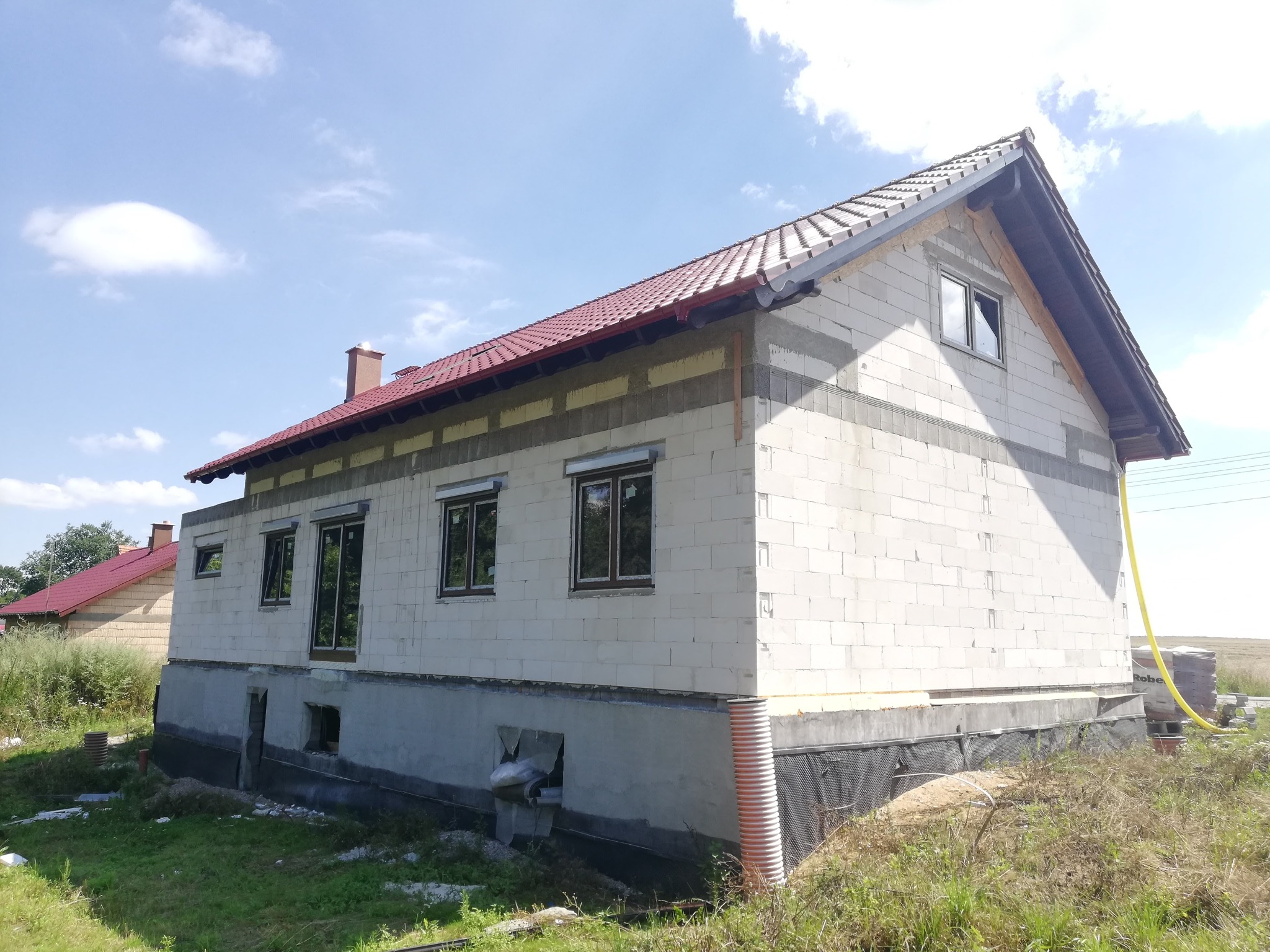 Surowy stan domu jednorodzinnego w trakcie budowy, widoczne okna z brązowymi ramami, ściany z białego bloczka, czerwony dach, żółty wąż ogrodowy i trawa na pierwszym planie.