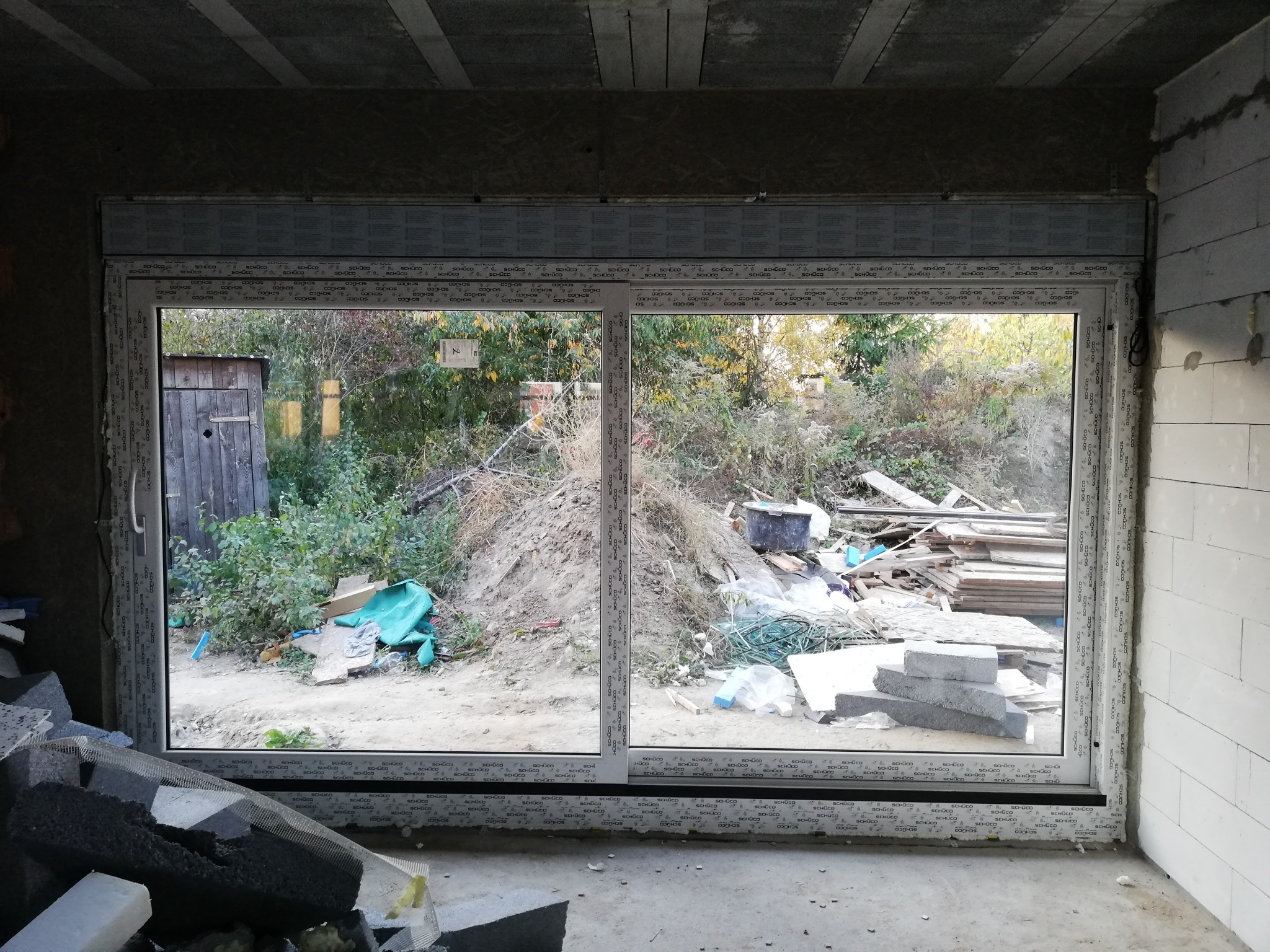 Nowe, białe okno PCV z widokiem na zaniedbany ogród z drewnianą szopą, widoczne bloki z betonu komórkowego i resztki materiałów budowlanych w surowym wnętrzu budynku.