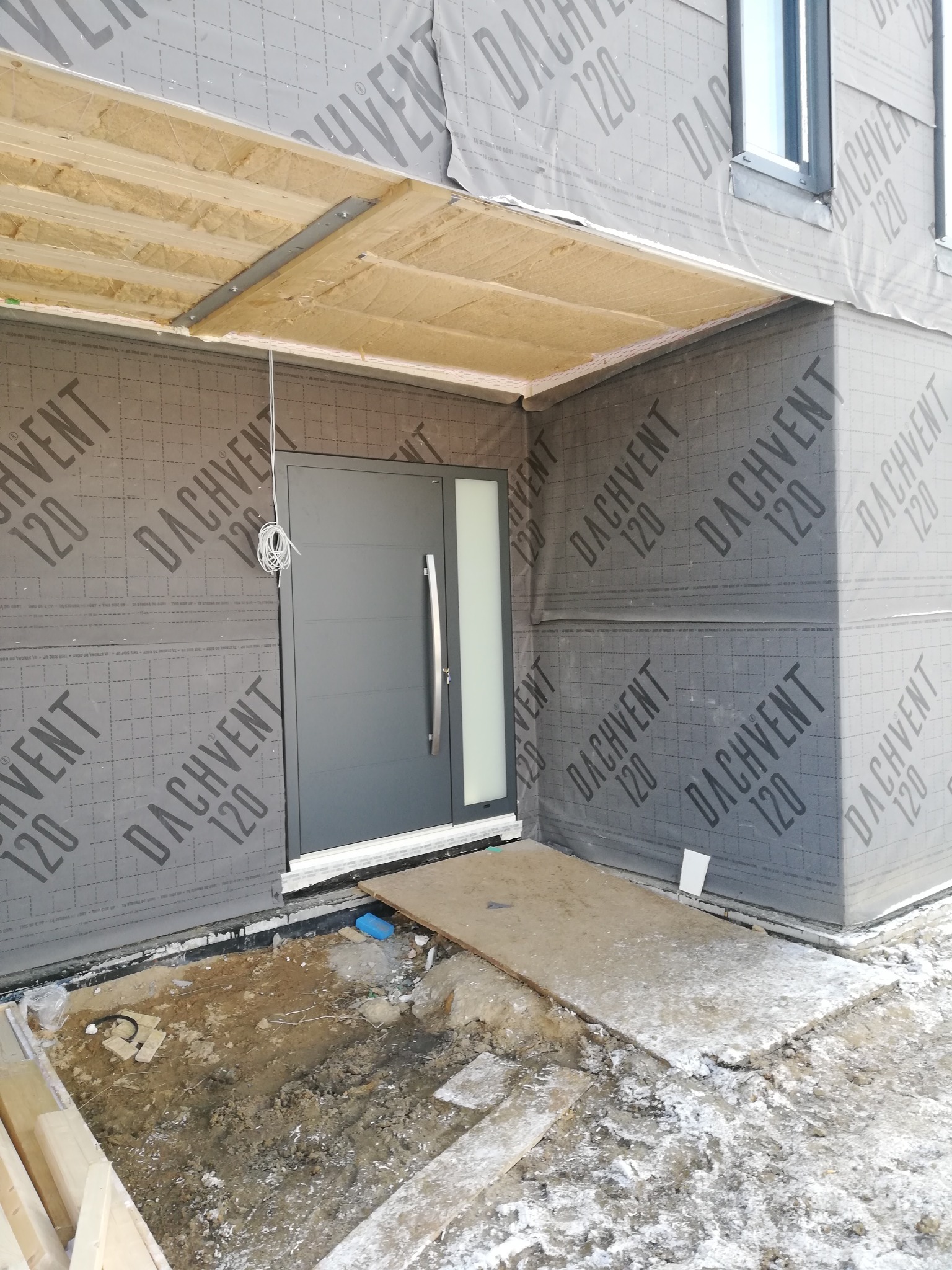 Szara, nowoczesna brama wejściowa z długim uchwytem, zamontowana w surowym stanie, z widocznym ociepleniem budynku i prowizorycznym wejściem z płyt.