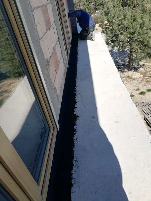 Pracownik na kolanach uszczelnia czarną masą połączenie okna z murem zewnętrznym budynku w słoneczny dzień.