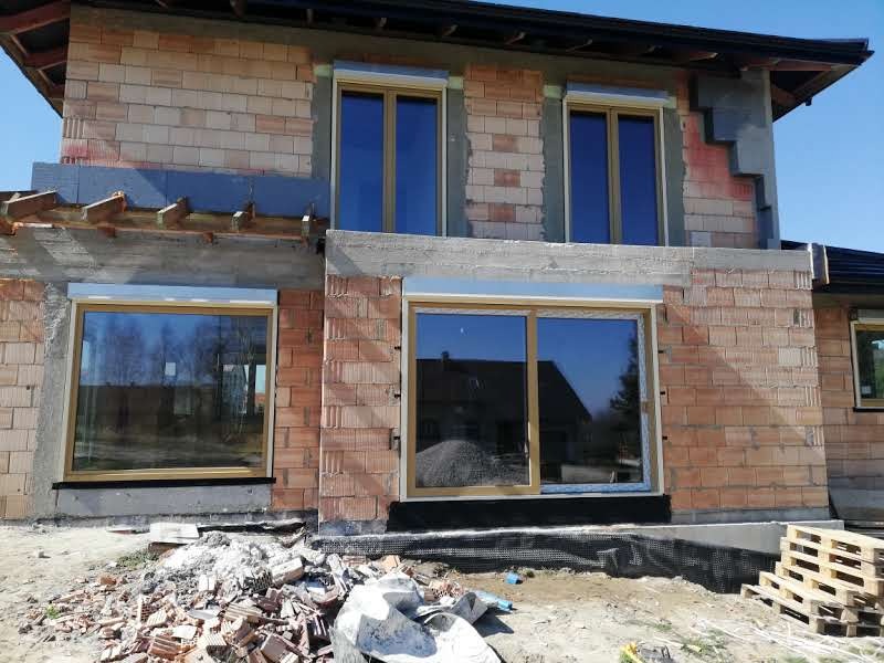 Nowo wstawione okna w surowym stanie, widoczne cegły i gruz na placu budowy, odblask budynku w szybie.