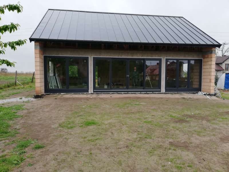 Surowy budynek z dużymi, przesuwnymi oknami w czarnych ramach, widoczne z zewnątrz na tle niezagospodarowanego terenu i częściowo wykończonych ścian z cegły.