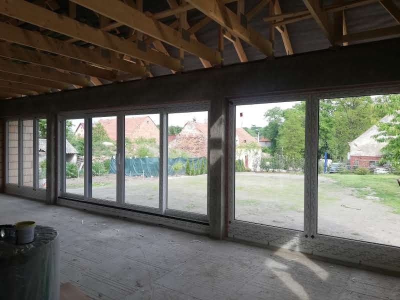 Surowy stan werandy z zamontowanymi oknami przesuwnymi z widokiem na ogród, widoczne drewniane krokwie dachu i niedokończona podłoga.