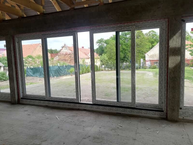 Wielkoformatowe przesuwne okno z profili PCV zamontowane w surowym stanie budynku z widokiem na trawnik i zabudowania gospodarcze.
