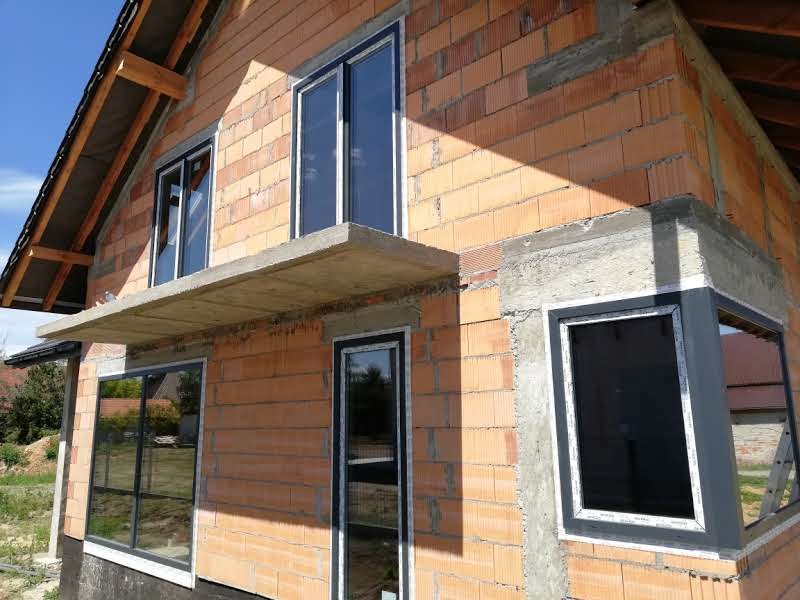 Surowy stan budynku w trakcie montażu okien z czarnymi ramami, widoczne elementy konstrukcyjne i cegły porotherm.