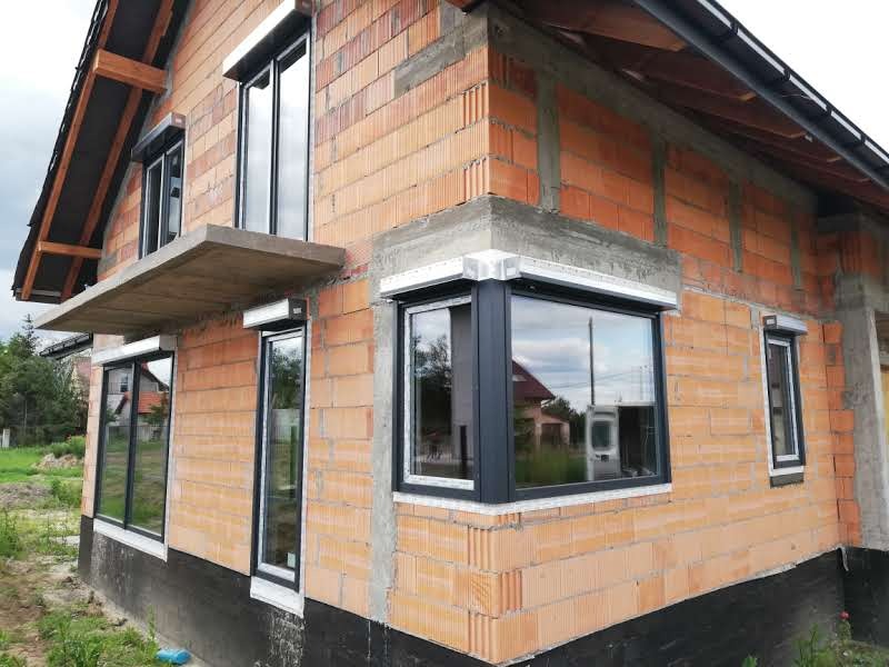 Niedokończony dom z zamontowanymi oknami z roletami zewnętrznymi, widoczne surowe ściany z cegły i betonowe nadproża.