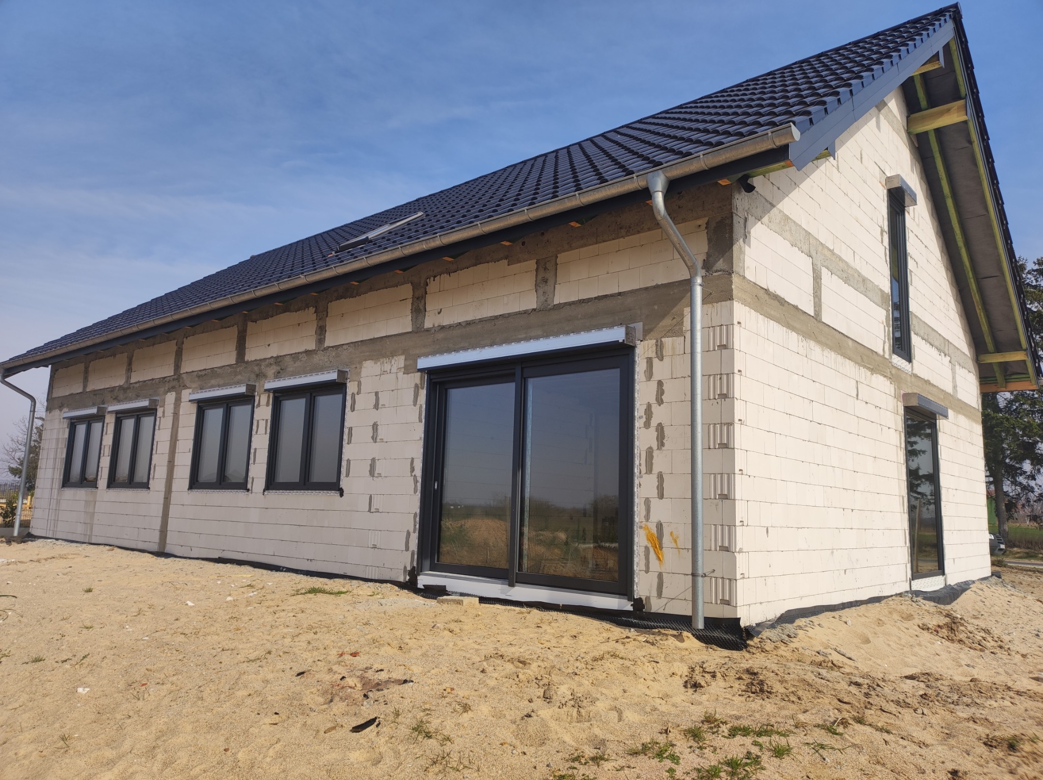 Nowo wybudowany dom jednorodzinny z czarnymi oknami i roletami zewnętrznymi, widoczne bloczki betonowe i niezagospodarowany teren wokół budynku.
