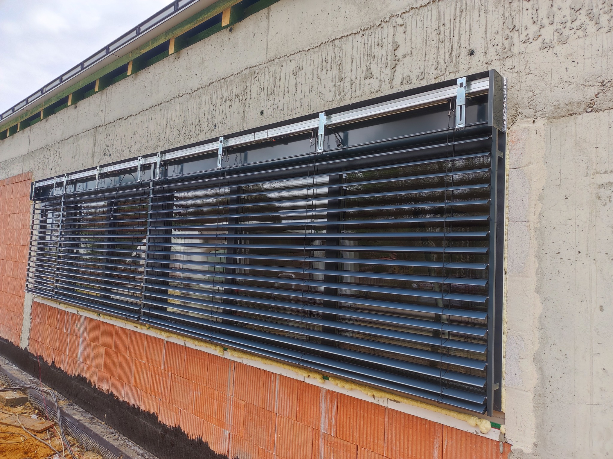 Zewnętrzne żaluzje fasadowe w kolorze antracytowym zamontowane w budynku w trakcie budowy, widoczne surowe ściany z betonu i cegły.