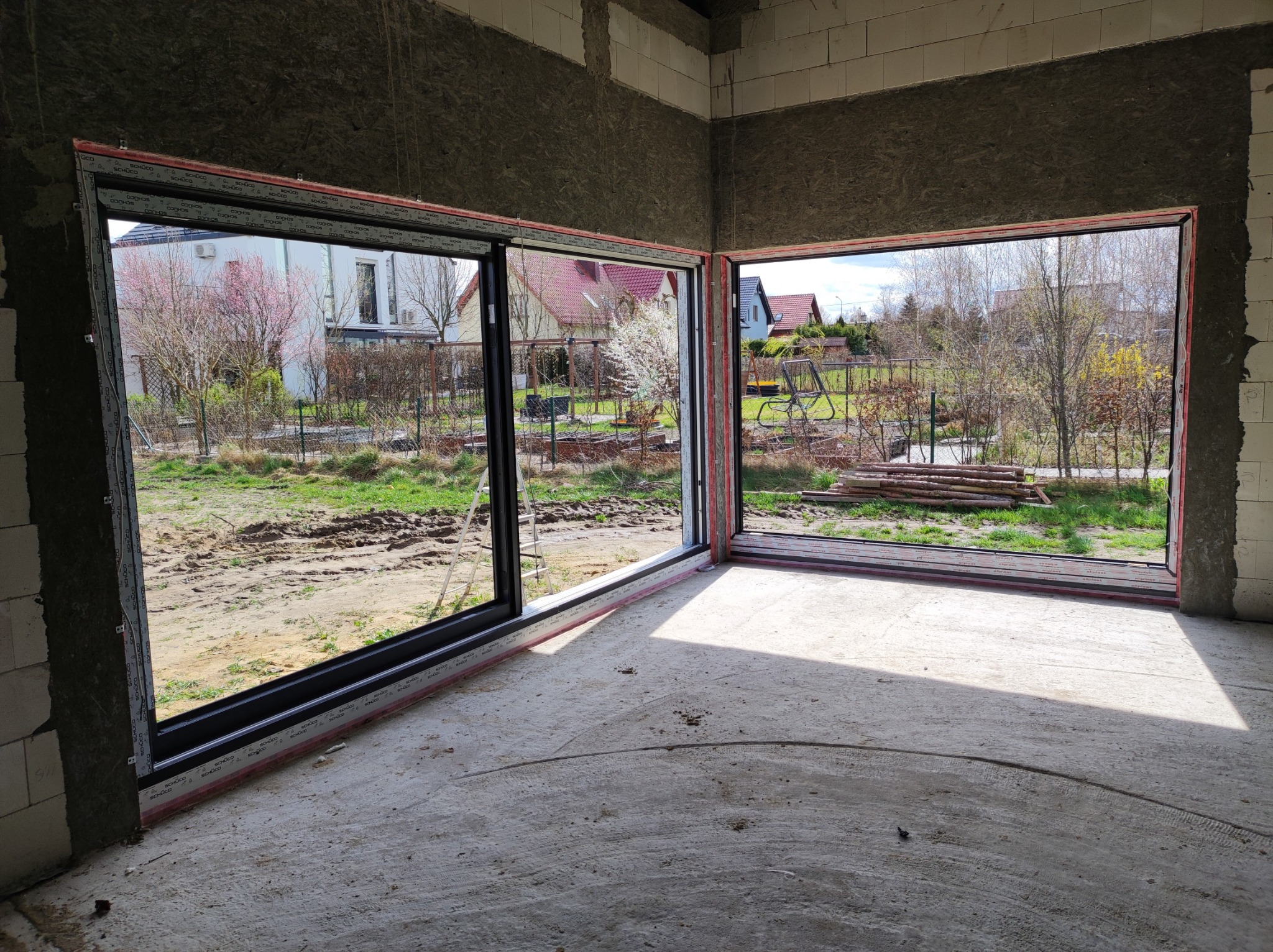 Surowy stan wnętrza z zamontowanymi dużymi przesuwnymi oknami z czarnymi ramami, widok na ogród z kwitnącymi drzewami, elementy konstrukcyjne ścian widoczne.