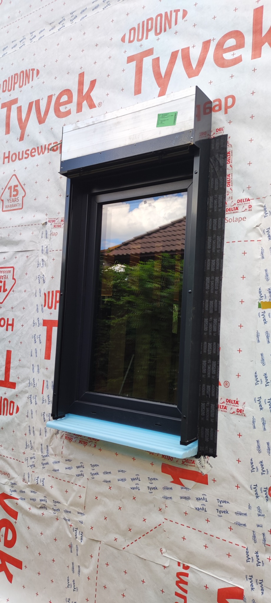 Ciemnoszare okno z roletą zewnętrzną w trakcie montażu, widoczne materiały izolacyjne i folia wiatroizolacyjna na ścianie.