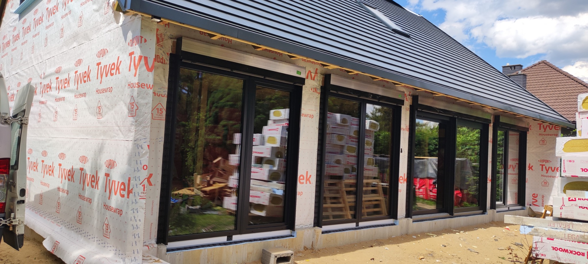 Budynek w trakcie budowy z zamontowanymi czarnymi oknami, ocieplony wełną mineralną i zabezpieczony folią Tyvek, widoczne materiały budowlane i rusztowania.