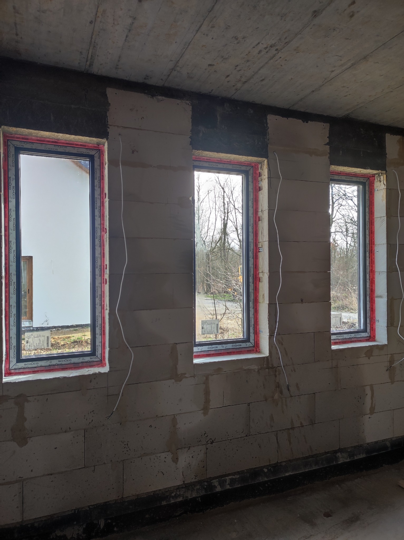 Surowy stan: Trzy okna z ramami Schüco, osadzone w ścianie z betonu komórkowego, widoczne kable elektryczne, brak wykończenia.