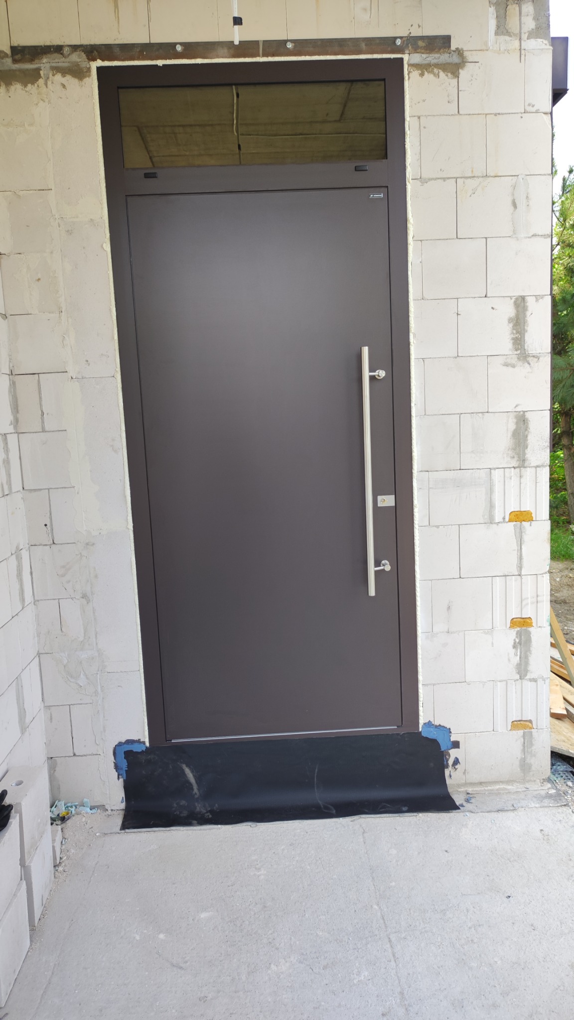 Brązowe drzwi wejściowe z długim, pionowym uchwytem zamontowane w surowym murze z betonu komórkowego, zabezpieczone folią na progu.