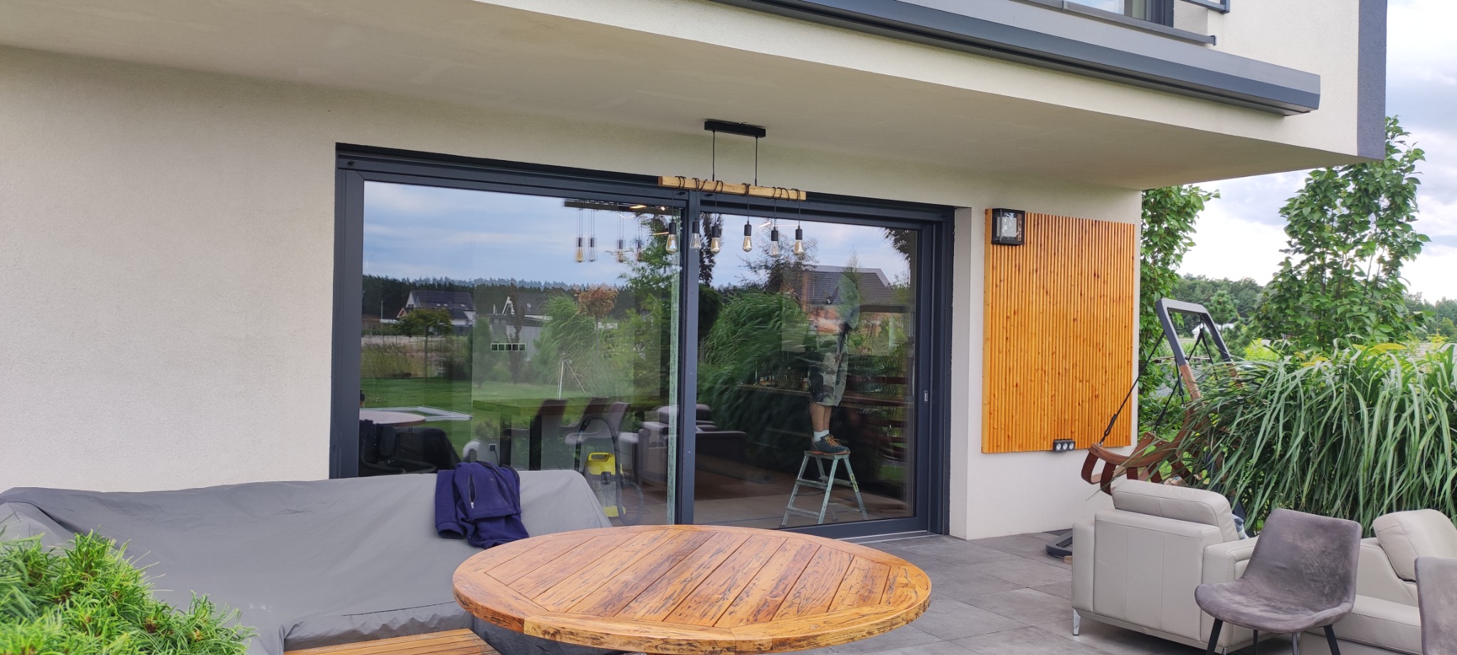 Duże przesuwne okno tarasowe z czarnymi ramami, odbijające postać na drabinie, widok z zewnątrz, drewniany stół na pierwszym planie.