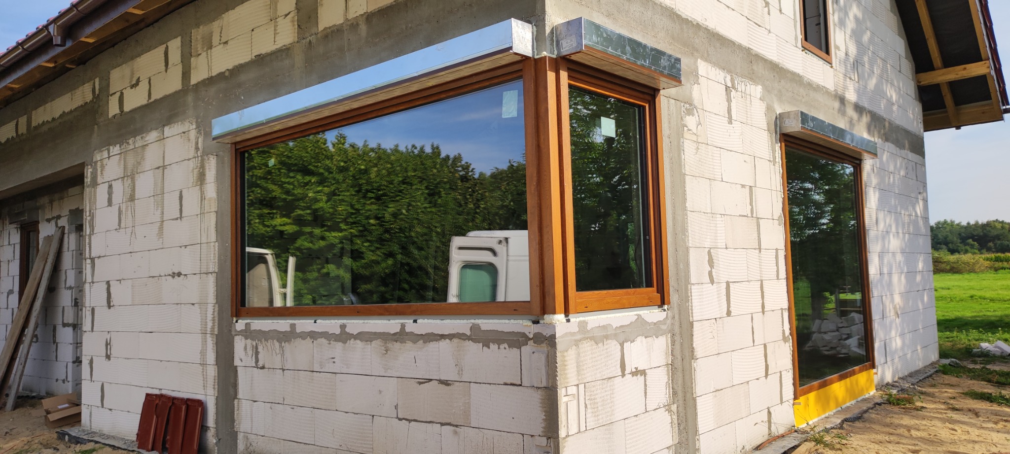 Nowo zamontowane okno PCV z brązową ramą i metalową osłoną zewnętrzną na surowej ścianie budynku w trakcie budowy, z widocznym odbiciem otoczenia.