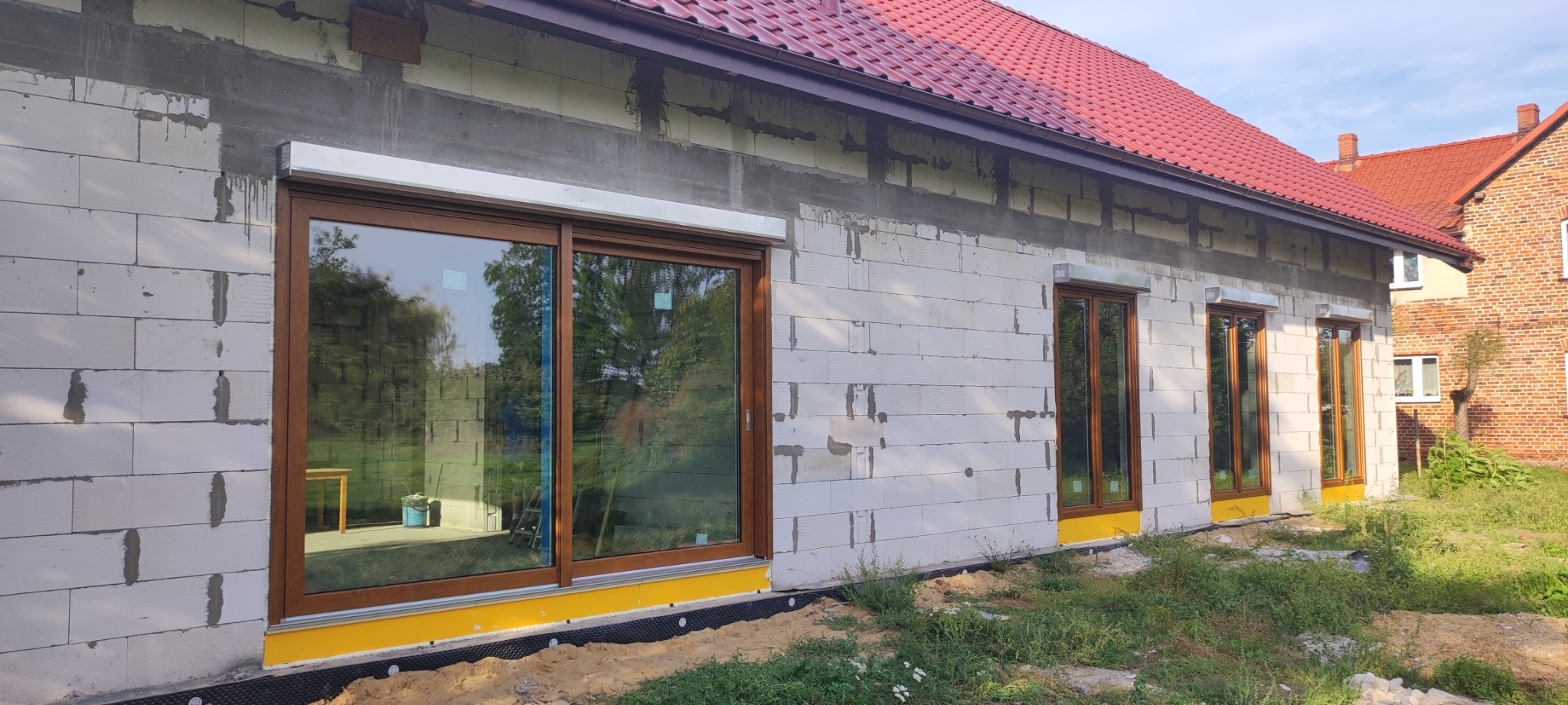 Montaż okien z brązowymi ramami w surowym budynku z białego bloczka, widoczne żółte zabezpieczenia i fragment darni.