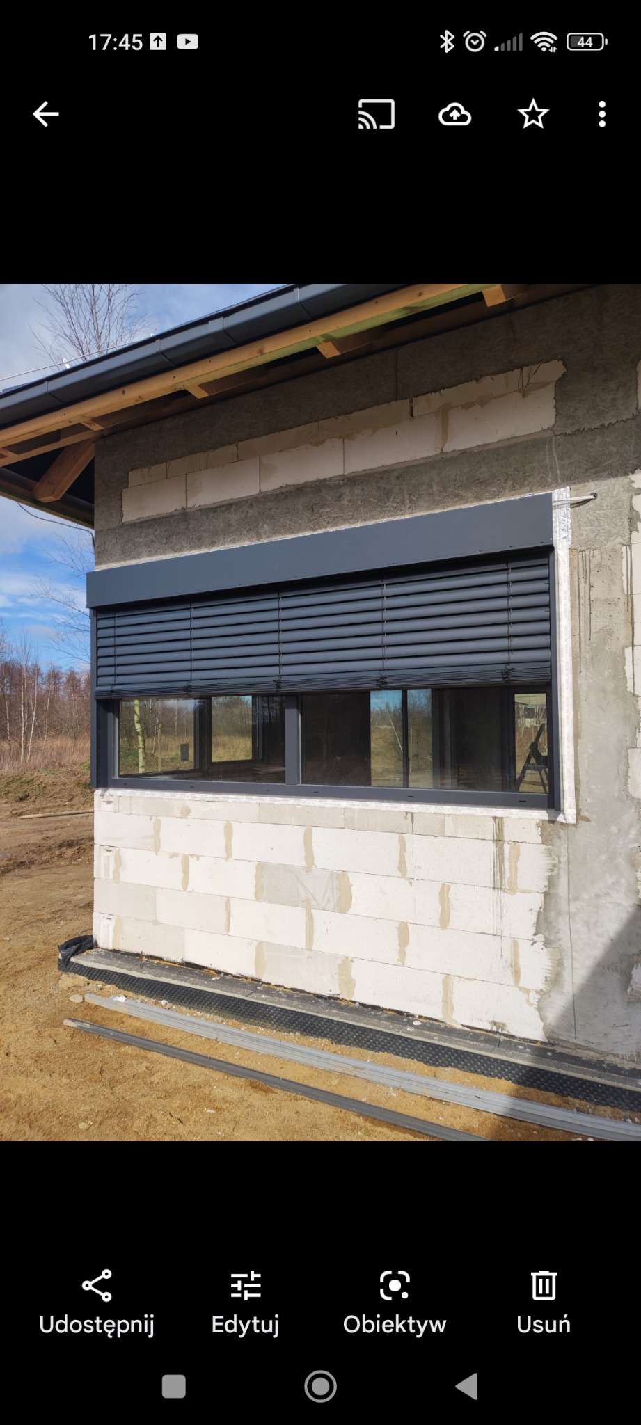 Elewacja surowego budynku z zamontowanymi zewnętrznymi roletami w kolorze grafitowym, widok na okno z zamkniętymi lamelami i fragment krajobrazu w tle.