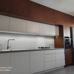 Glam House - Nowoczesna kuchnia z białymi, matowymi frontami szafek i blatem w kolorze szarości, uzupełniona drewnianą zabudową sięgającą sufitu, z oświetleniem LED pod górnymi szafkami i teksturowanymi...