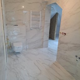 Glam House - Wykończona łazienka z białymi marmurkowymi płytkami, wisząca toaleta z folią ochronną, grzejnik drabinkowy i nietypowe wejście do kabiny prysznicowej z ukośną ścianą.