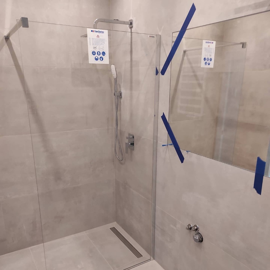 Nowoczesna kabina prysznicowa typu walk-in z odpływem liniowym, zabezpieczona niebieską taśmą malarską na szklanych elementach, zamontowana na tle płytek w odcieniach szarości, z widoczną armaturą.